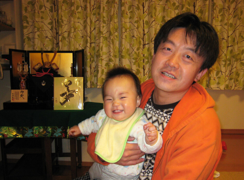 鎧兜を背景に、満面の笑みでカメラ目線の息子さんを胸に抱き、カメラに微笑む鈴木先生